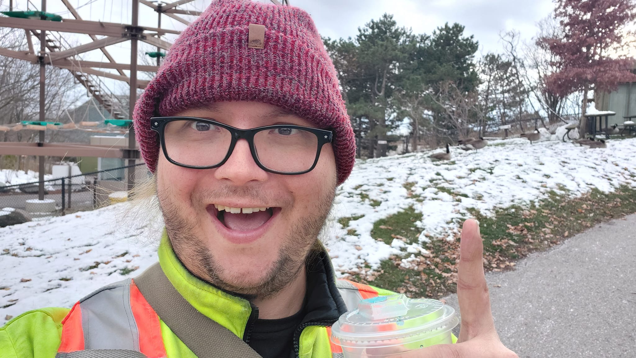 A man wearing a red toque and a high visibility winter coat points at geese behind him while holding a blue slushie